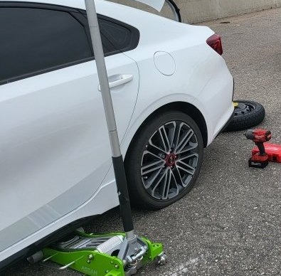 Tire change: a white car on a floor jack, the spare tire and an impact wrench on the asphalt